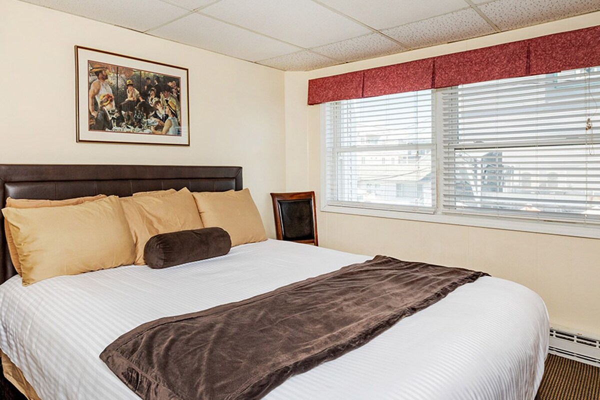 A neatly made bed with a headboard, a bedside table with a lamp, a framed picture on the wall, and a window with blinds.
