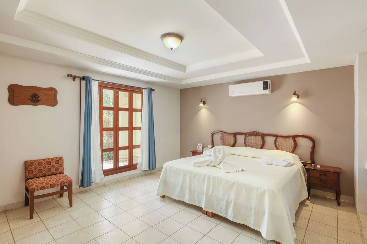Habitación de hotel con cama, una silla, mesita de noche, equipo de aire acondicionado y ventana con cortinas.