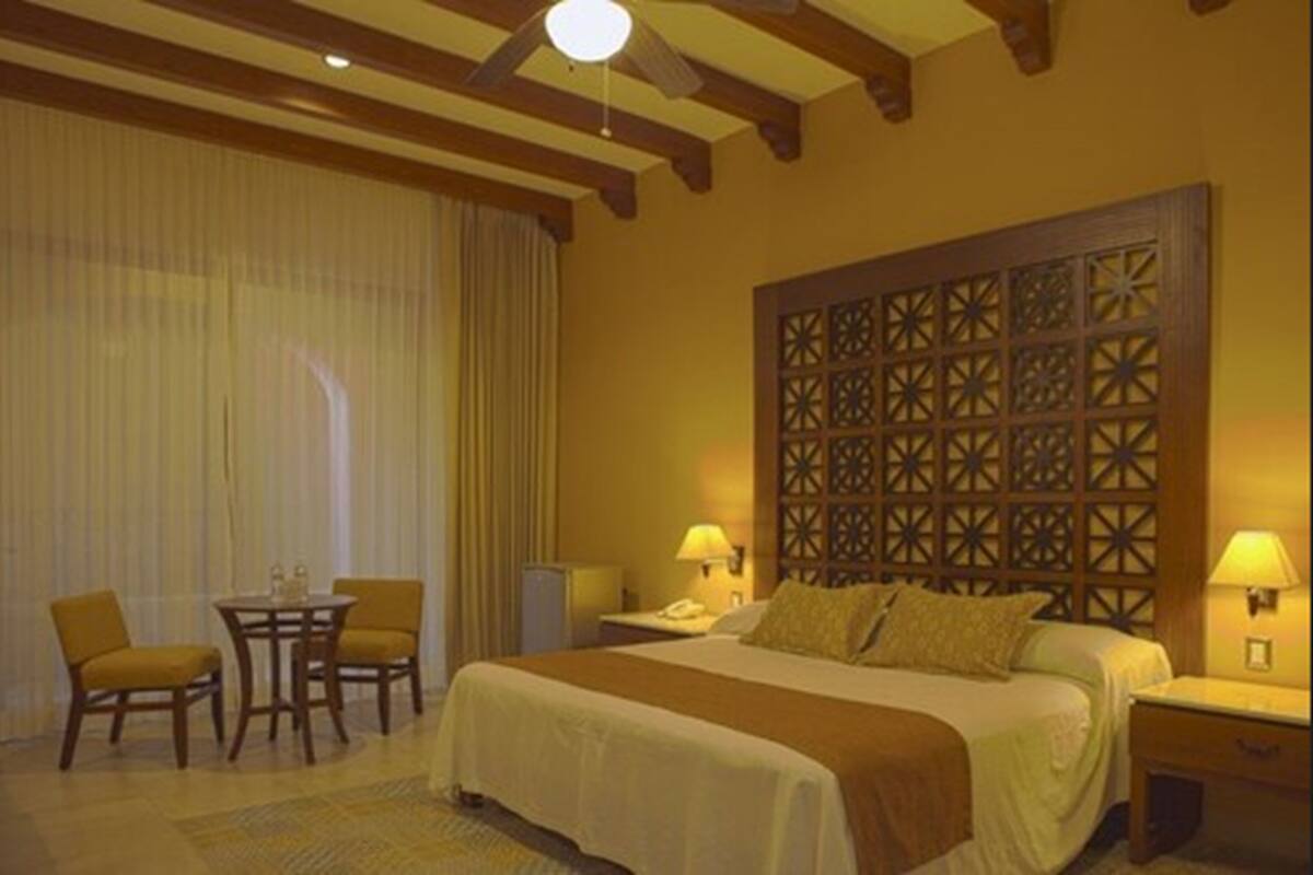 Habitación de hotel con una cama grande, cabecera de madera, una mesita pequeña con dos sillas y una mesita de noche con lámpara.