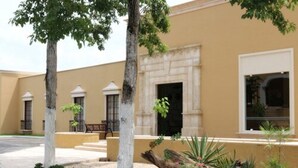 Un bâtiment à la façade beige, avec une entrée centrale dotée d’une porte blanche et un petit jardin agrémenté d’arbres et de plantes.
