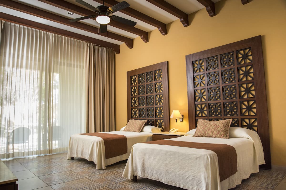 Habitación de hotel con dos camas, un ventanal con cortinas, un ventilador de techo y un panel de madera decorativo en la pared.