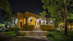 Une maison bien éclairée, à deux étages, avec un escalier menant à l’entrée, entourée d’une végétation luxuriante et de palmiers.