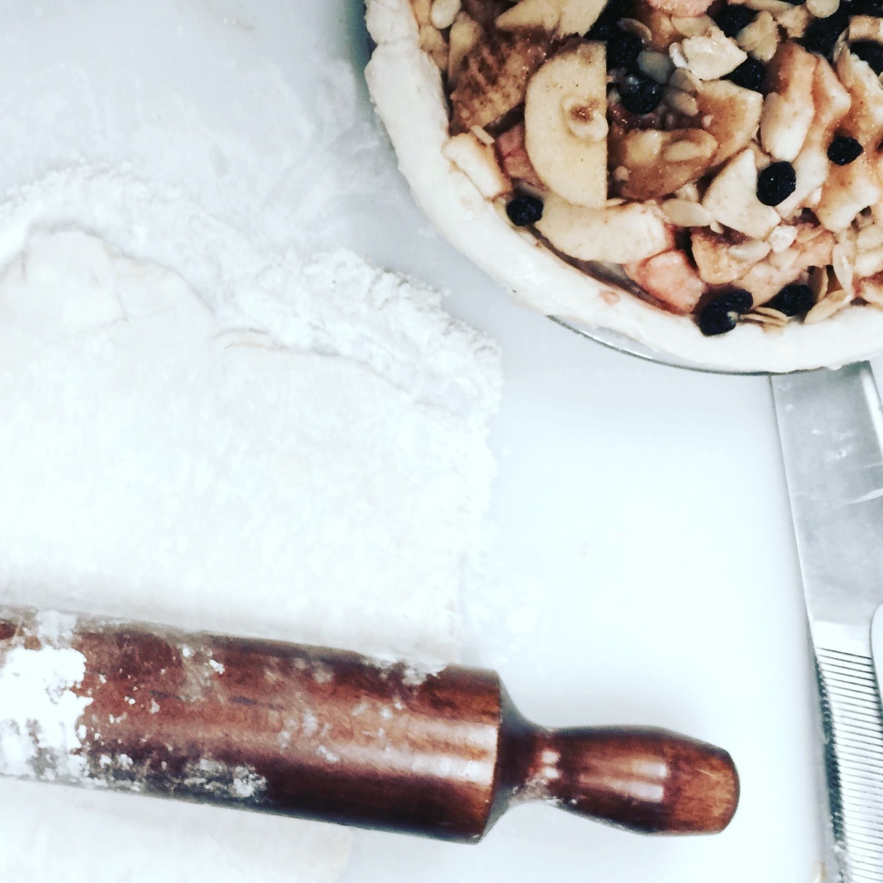 A kitchen counter with a rolling pin, a bowl of flour, and a partially prepared pie with sliced apples and blackberries.