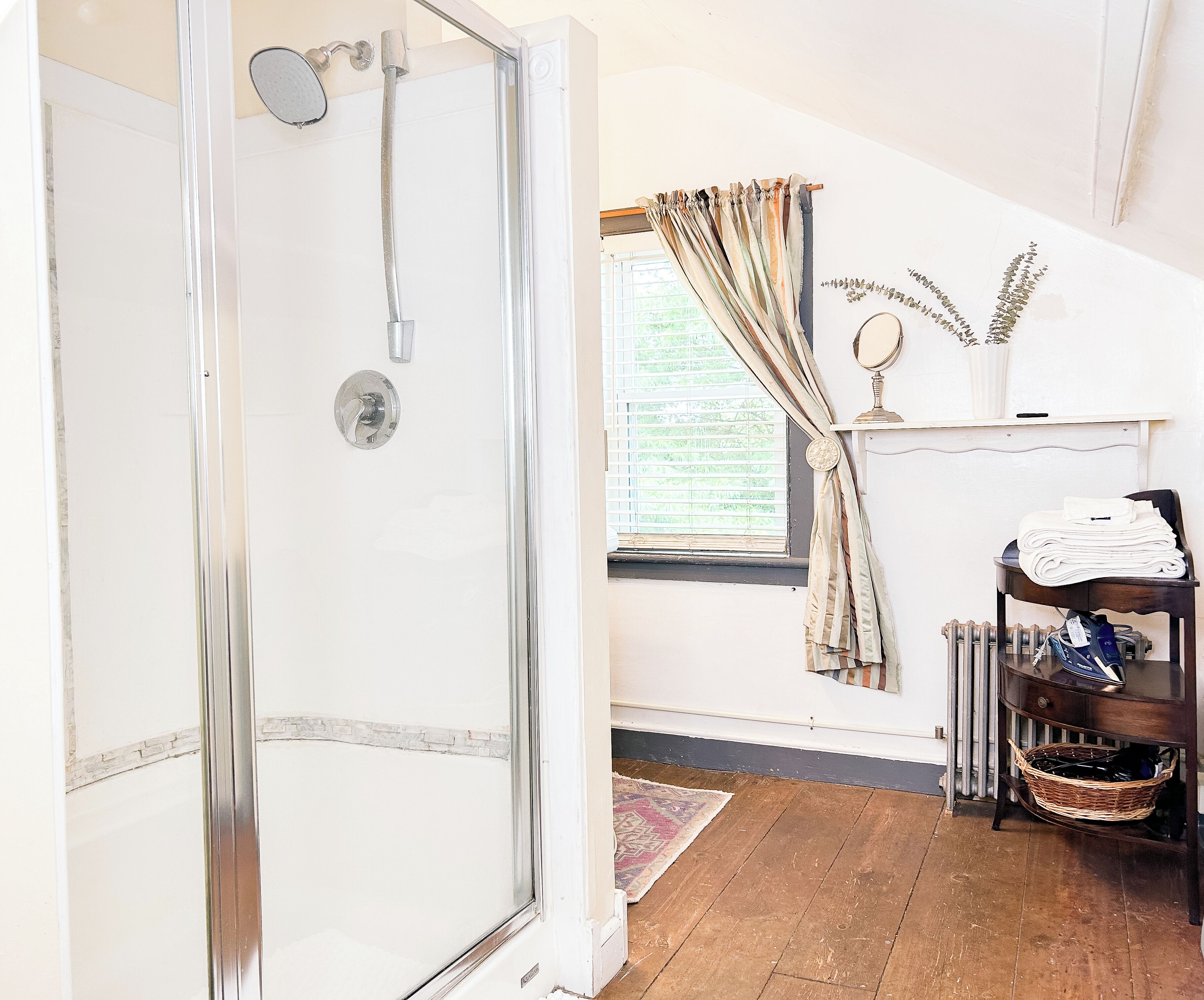 Une salle de bain avec une porte de douche en verre, une fenêtre avec des rideaux, un radiateur et une étagère avec des serviettes pliées.