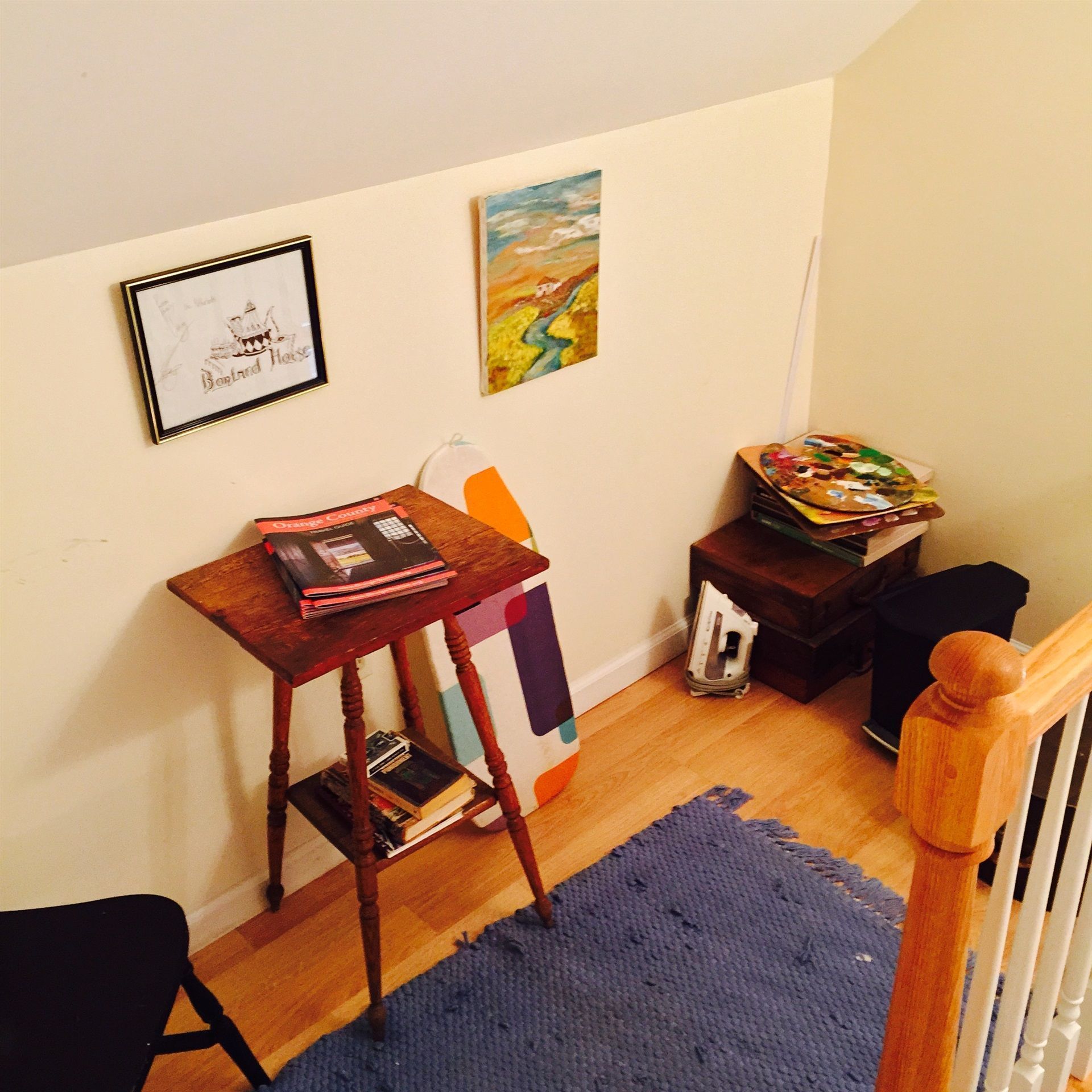 Una habitación con una mesa de madera, un cuadro enmarcado, un cuadro colorido y un taburete pequeño.