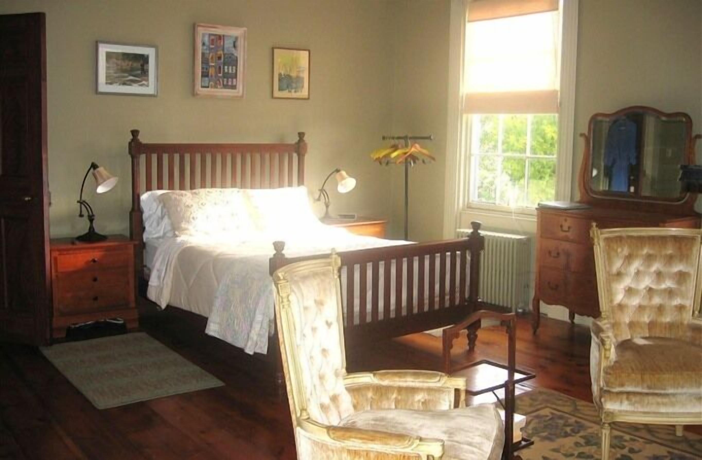 Un dormitorio con cama de madera, dos mesitas de noche con lámparas, un aparador, una silla y una ventana con vistas a la vegetación.