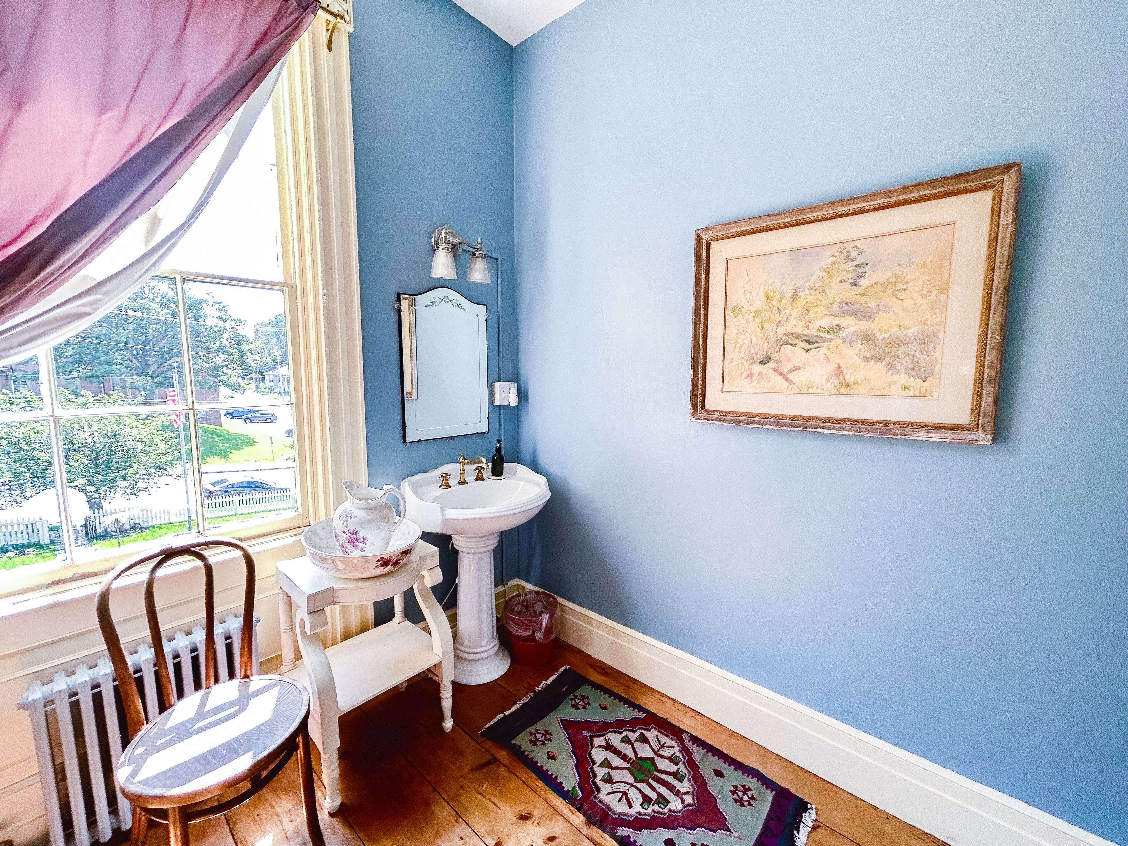 A bathroom with a white pedestal sink, a mirror, and a framed landscape painting on the wall.