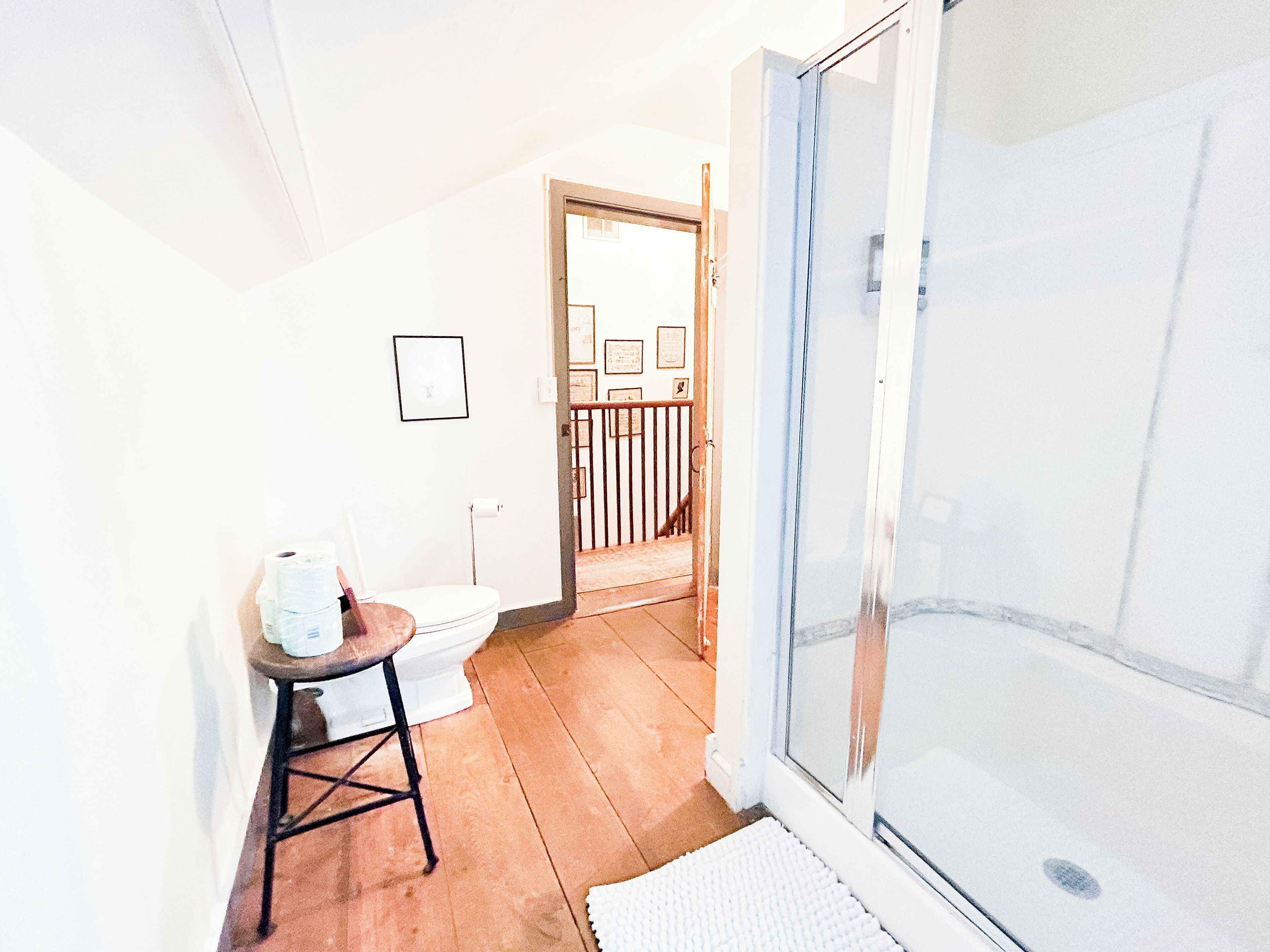 A bathroom with a toilet, a small stool, a shower with a glass door, and a wooden floor.