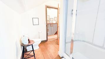 A bathroom with a toilet, a small stool, a shower with a glass door, and a wooden floor.