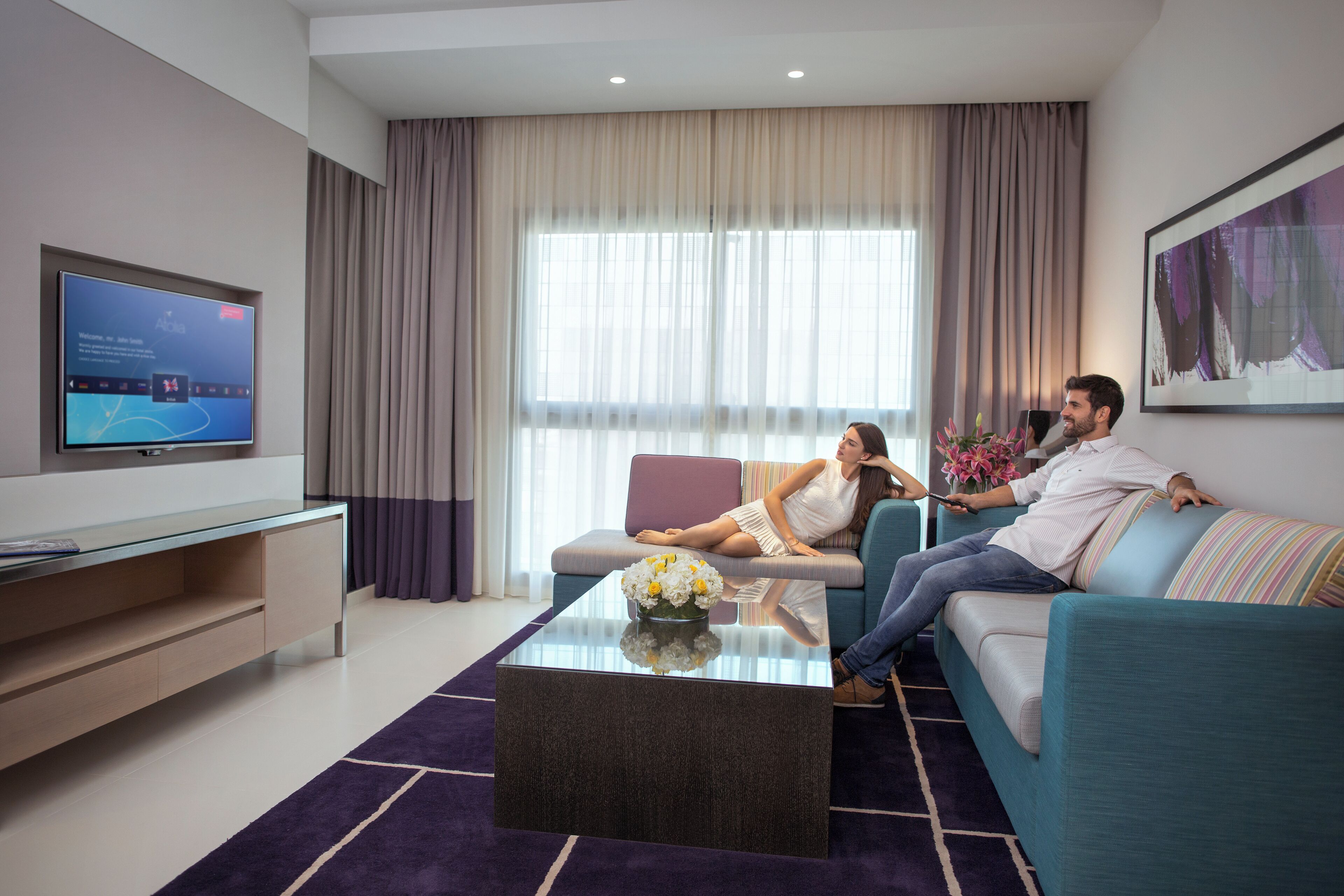 Ett modernt vardagsrum med en platt-TV, en soffa, ett soffbord och ett stort fönster med gardiner.