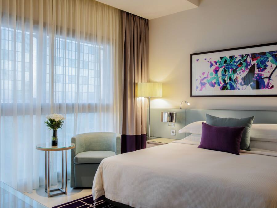A hotel room with a bed, a chair, a small table with a vase of flowers, a lamp, and a painting on the wall.