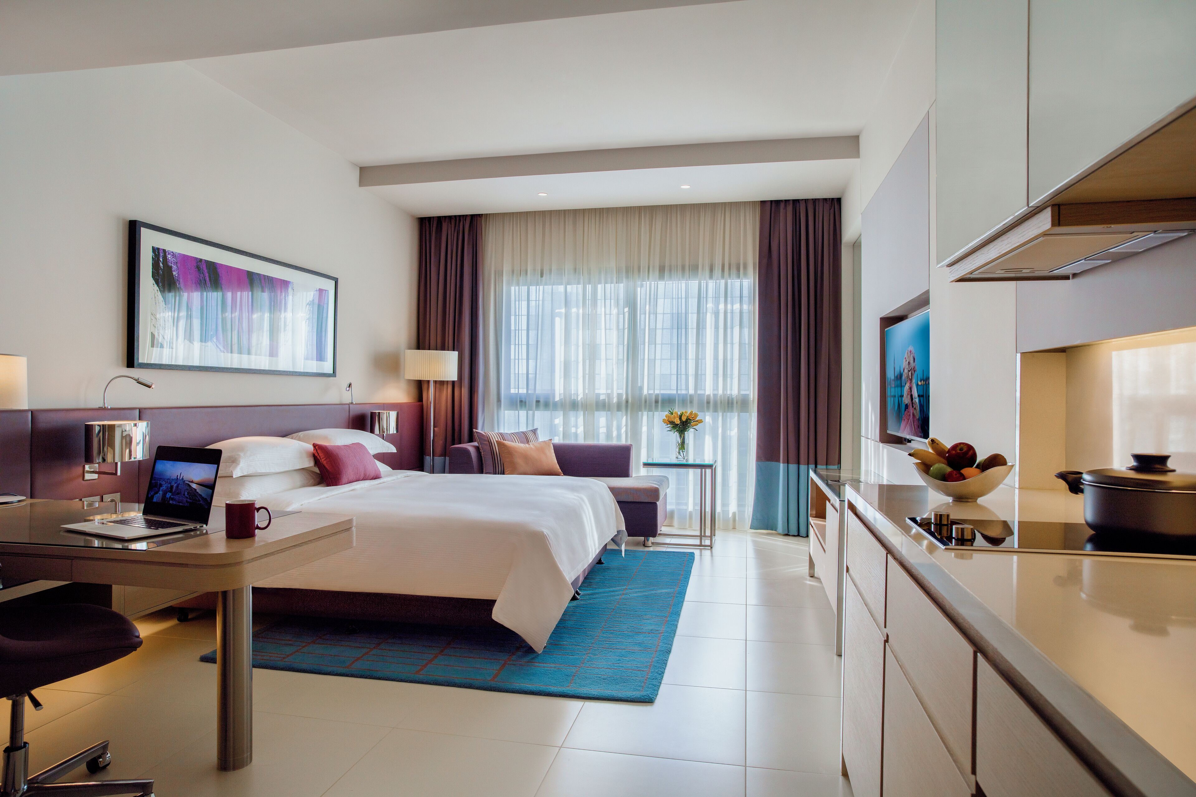 A modern hotel room with a bed, desk, laptop, and a dining area with a bowl of fruit.