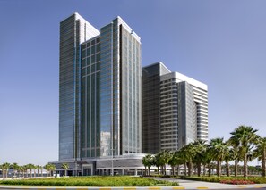 Un immeuble moderne de grande hauteur, doté d’une façade en verre, entouré de palmiers et agrémenté d’un espace paysager.