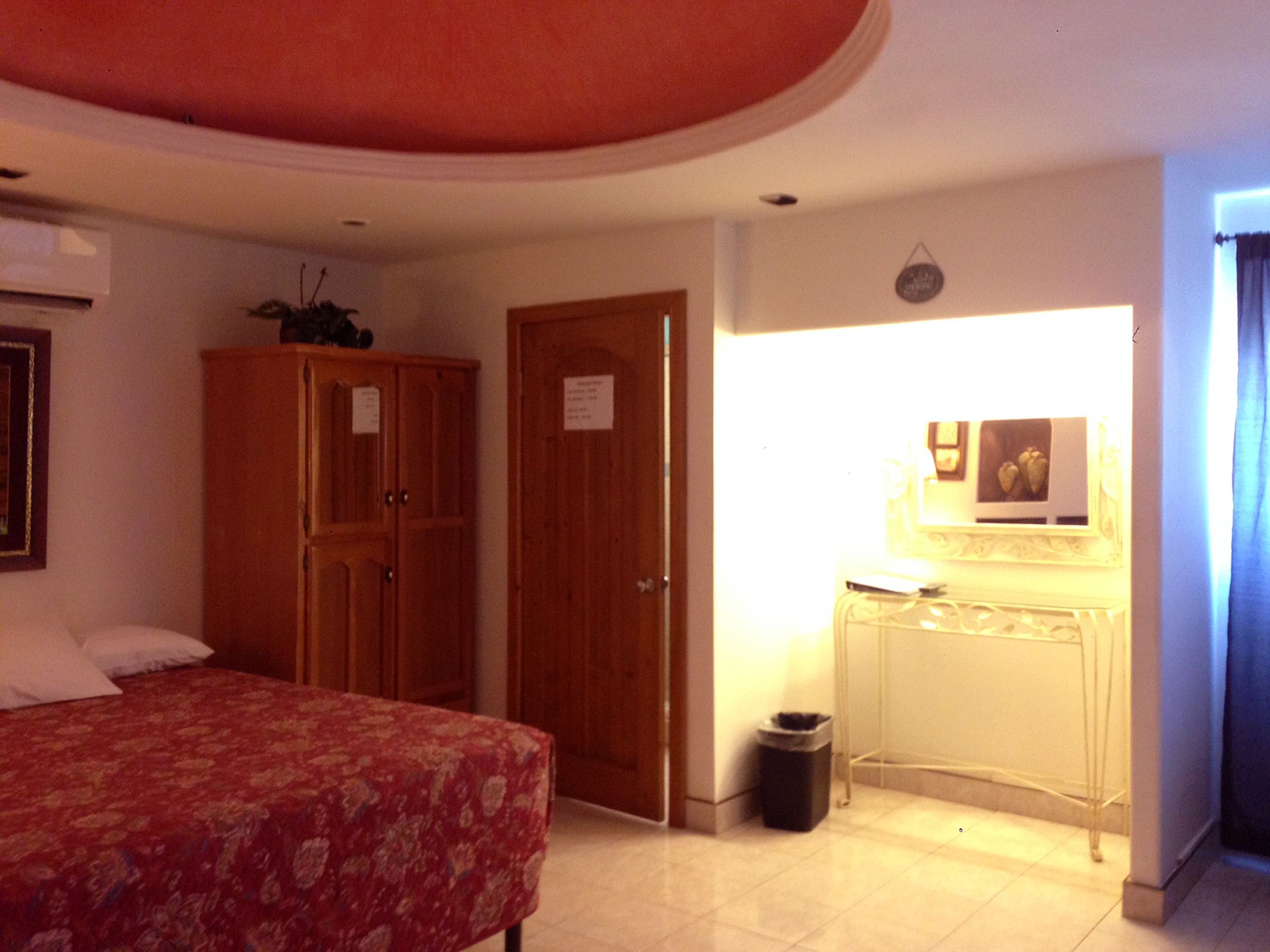 Une chambre d’hôtel avec un lit, une armoire en bois, un bureau avec une lampe et une poubelle.