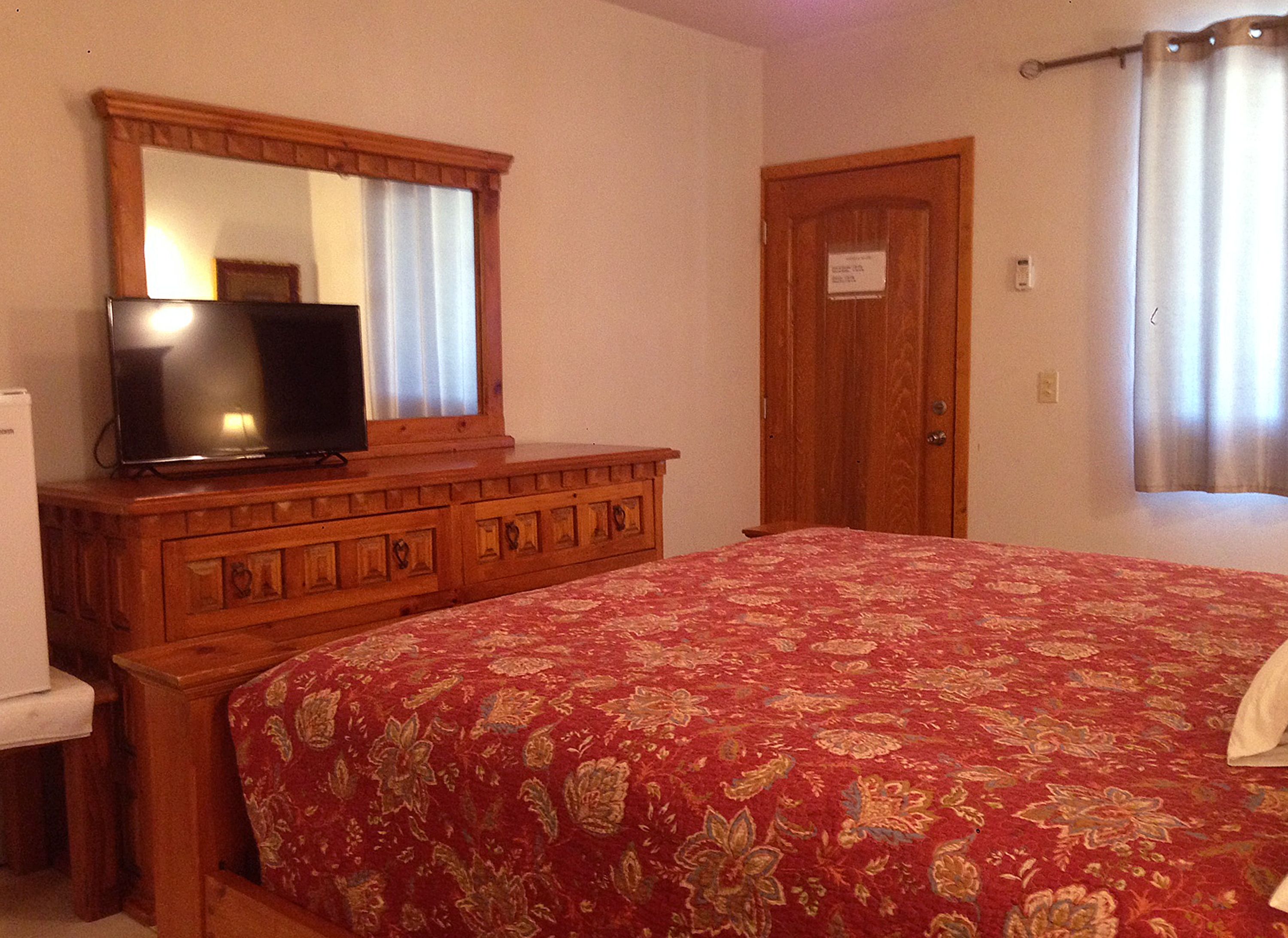 Un dormitorio con un aparador de madera, televisor, cama con colcha roja estampada, una puerta y una ventana con cortinas.