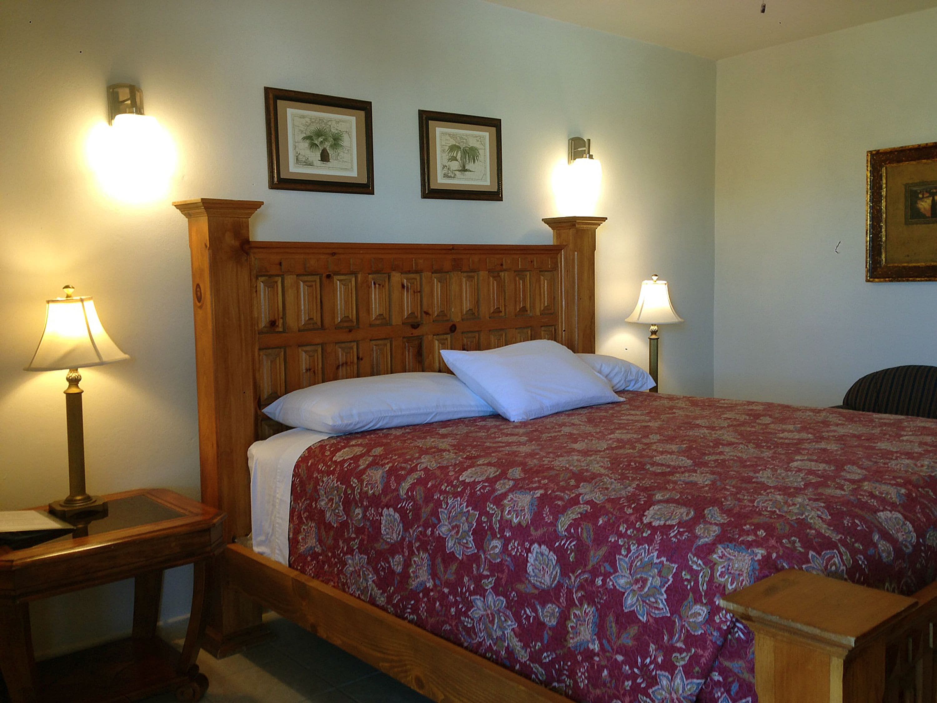 A bedroom with a wooden headboard, floral bedspread, two bedside lamps, and framed artwork on the wall.
