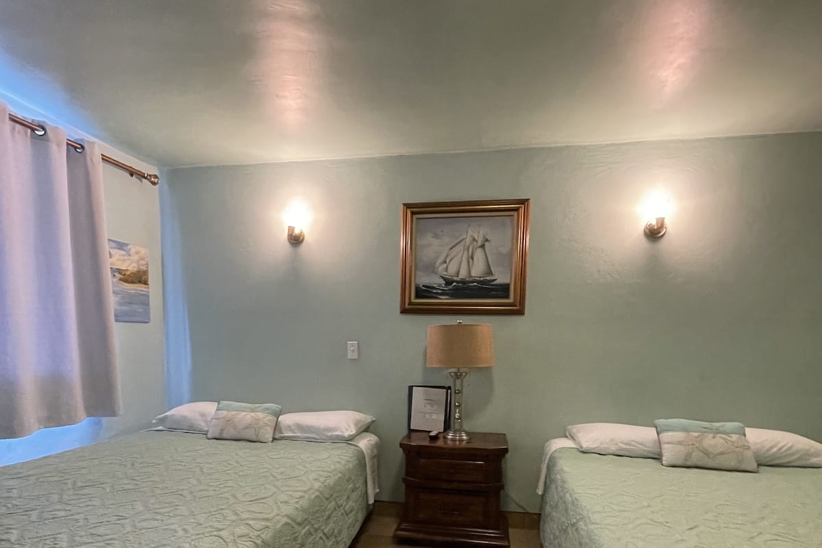 Ein Zimmer mit zwei Betten, einem Nachttisch mit Lampe, einem gerahmten Bild an der Wand und einem Fenster mit Vorhängen.