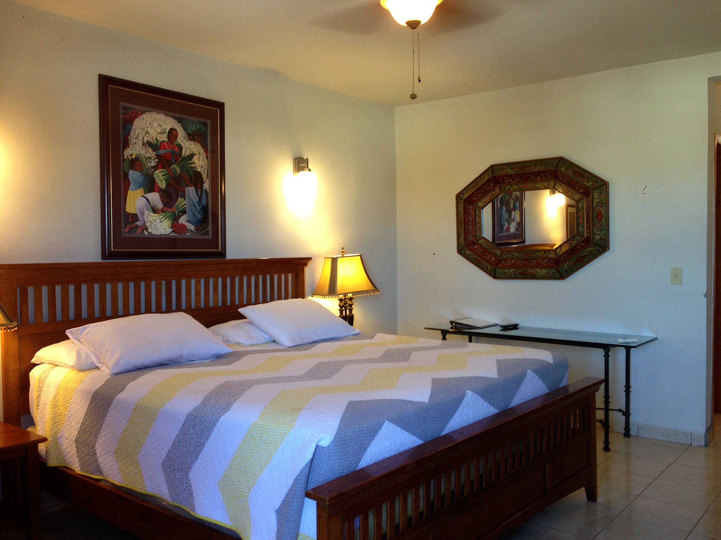Una cama bien hecha con una manta a rayas, una lámpara de noche, un cuadro enmarcado en la pared y un espejo con marco decorativo.