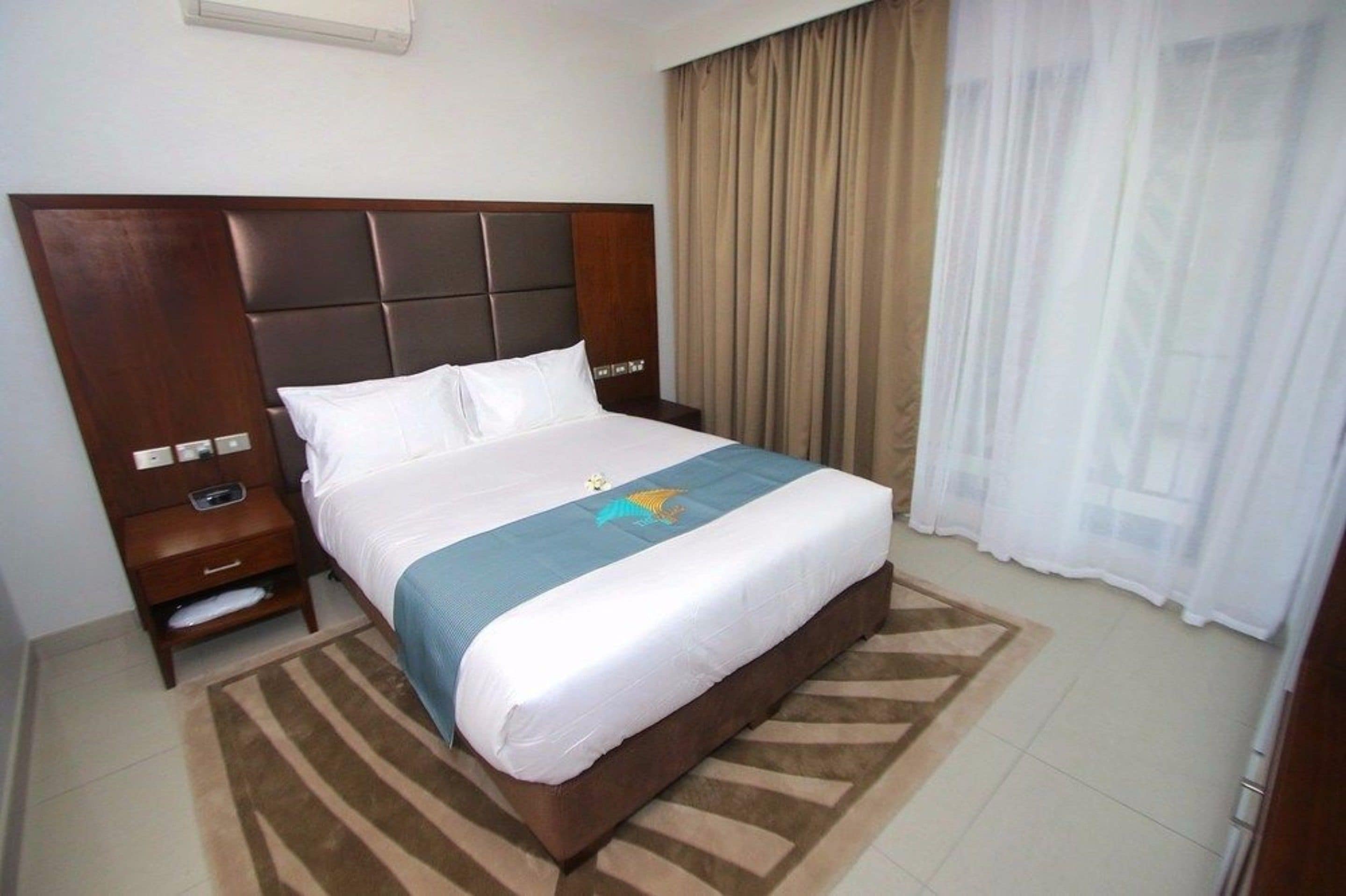 Une chambre d’hôtel avec un grand lit, une tête de lit en bois, une table de chevet et une commode, ainsi qu’une fenêtre avec des rideaux.