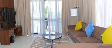 Una sala de estar moderna con un sofá, una mesa de centro, un mueble para televisión y ventanas grandes con cortinas.