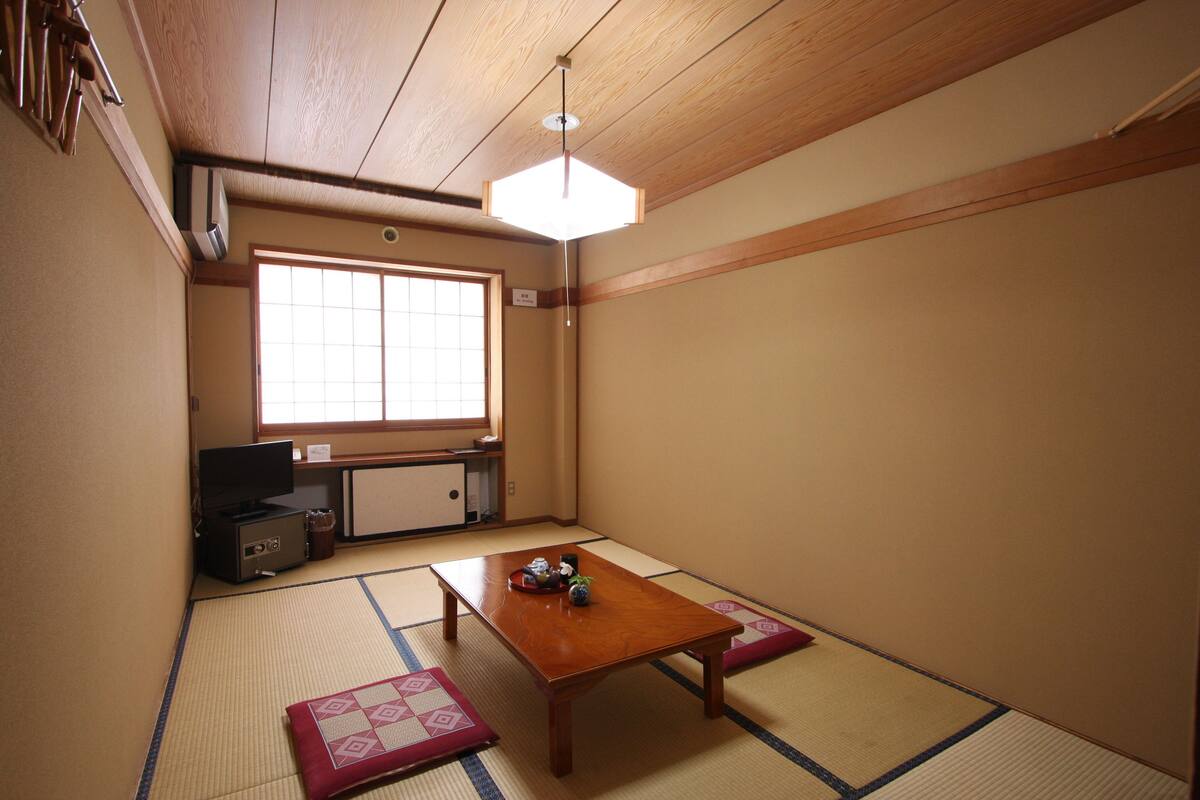 Ein traditioneller japanischer Raum mit Tatami-Bodenbelag, einem niedrigen Holztisch, einem Fernseher und einem Fenster, das natürliches Licht hereinlässt.