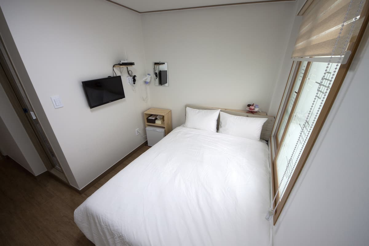 A bedroom with a bed, a TV mounted on the wall, a bedside table, and a window with blinds.