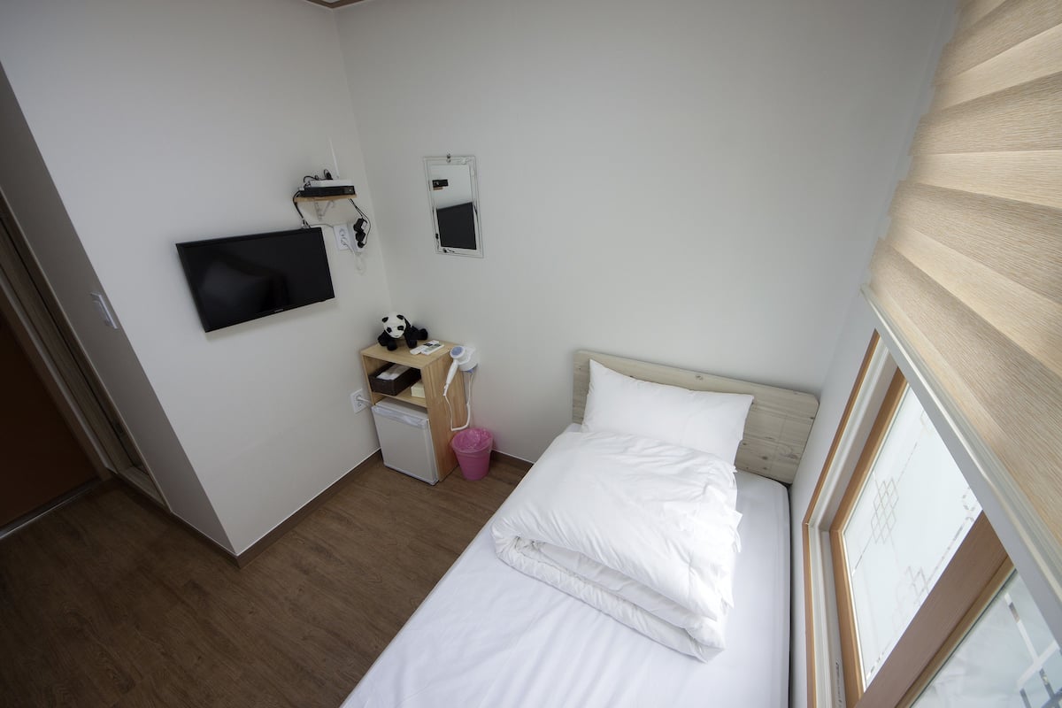 A hotel room with a bed, a TV, a small desk, and a window with blinds.