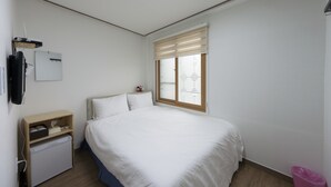 A bedroom with a bed, a small desk, a refrigerator, a window with blinds, and a ceiling light. - Charim Guesthouse (Seoul)