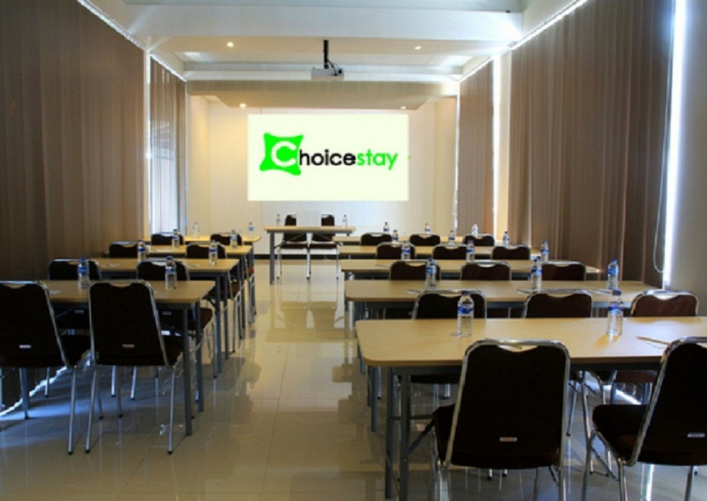 Una sala de conferències amb files de taules i cadires, una pantalla de projector que mostra 'ChoiceStay' i un atril per a presentacions.