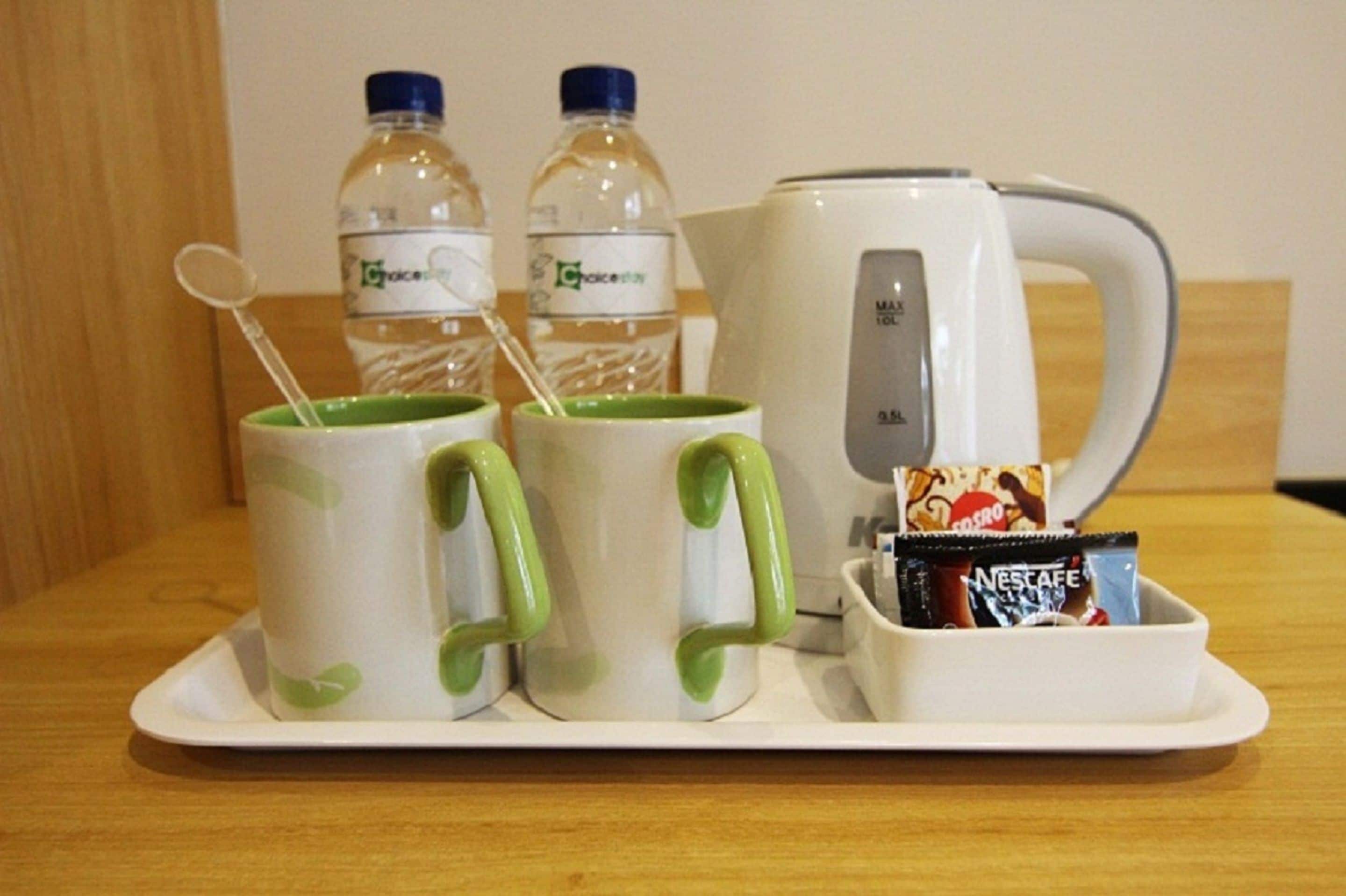 Una bandeja con dos tazas de mango verde, dos botellas de agua, una tetera eléctrica blanca y un recipiente con un sobre de Nescafé.
