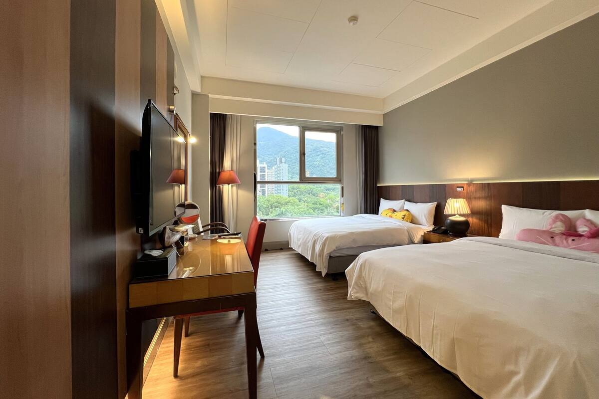 A hotel room with two beds, a desk, a television, and a window with a view of buildings and greenery.