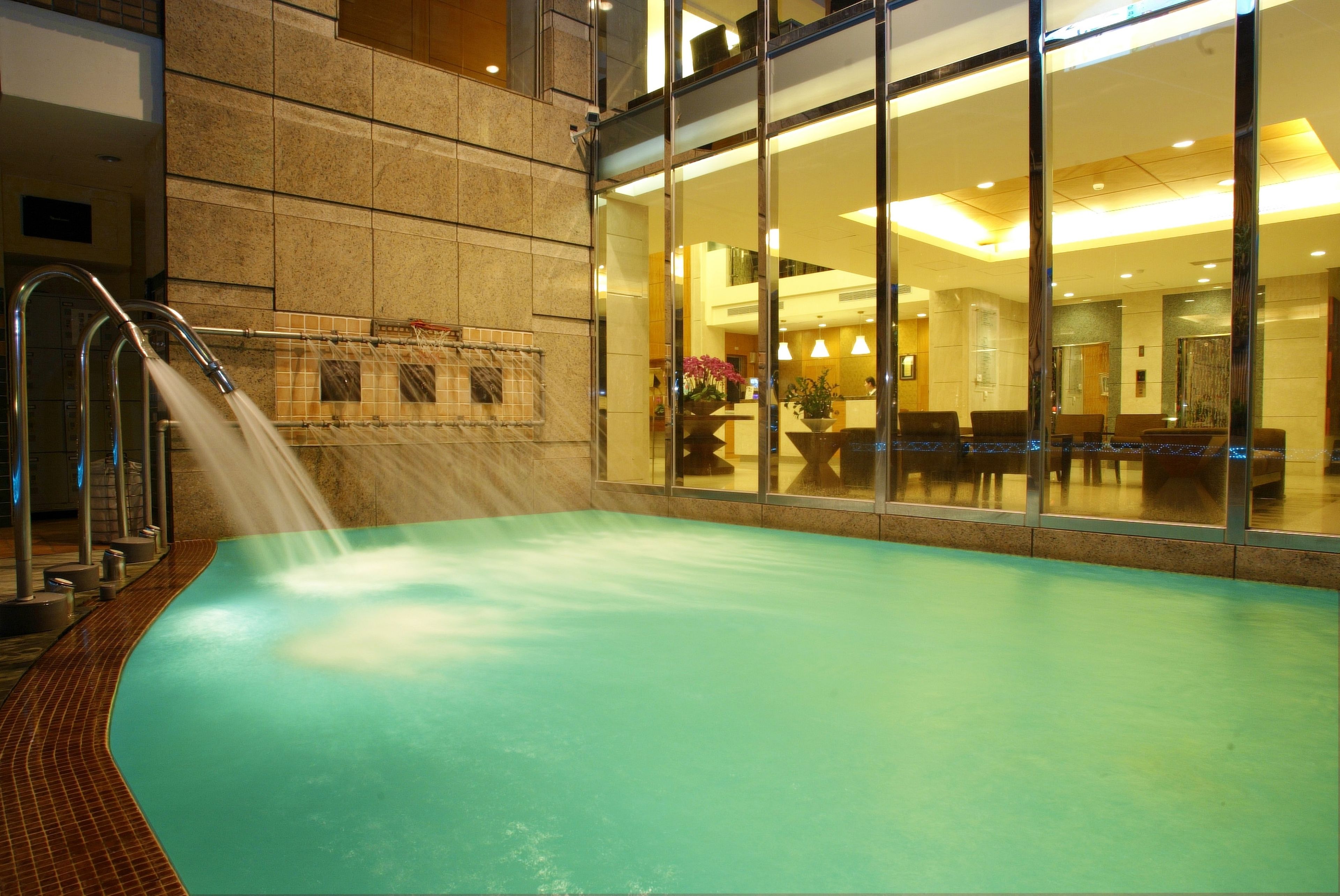 Una piscina interior con suelo de baldosas, un elemento acuático y un área de recepción visible a través de puertas de vidrio.