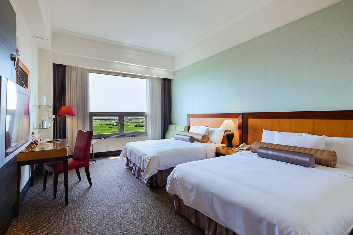 A hotel room with two beds, a desk with a chair, a lamp, and a view of the outdoors.