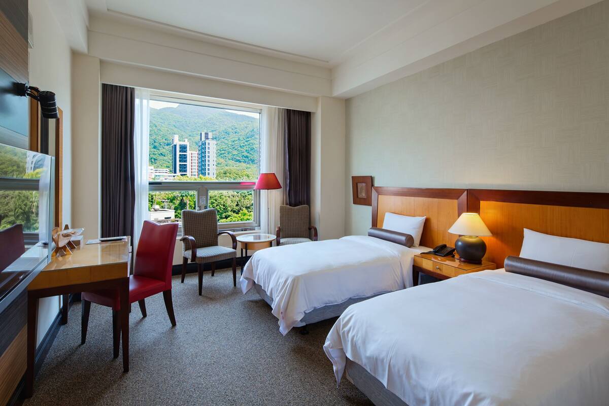 A hotel room with two beds, a desk, a chair, a small table, and a window with a view of buildings and greenery.