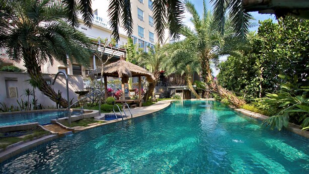 A swimming pool with a thatched-roof structure, surrounded by lush greenery and a multi-story building in the background. - Hotel Les Champs Jiaosi (Jiaoxi)