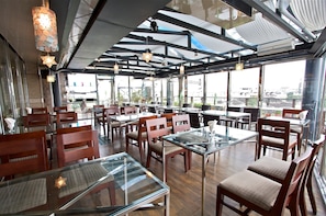 A restaurant with glass tables and wooden chairs, large windows, and a high ceiling with exposed beams.