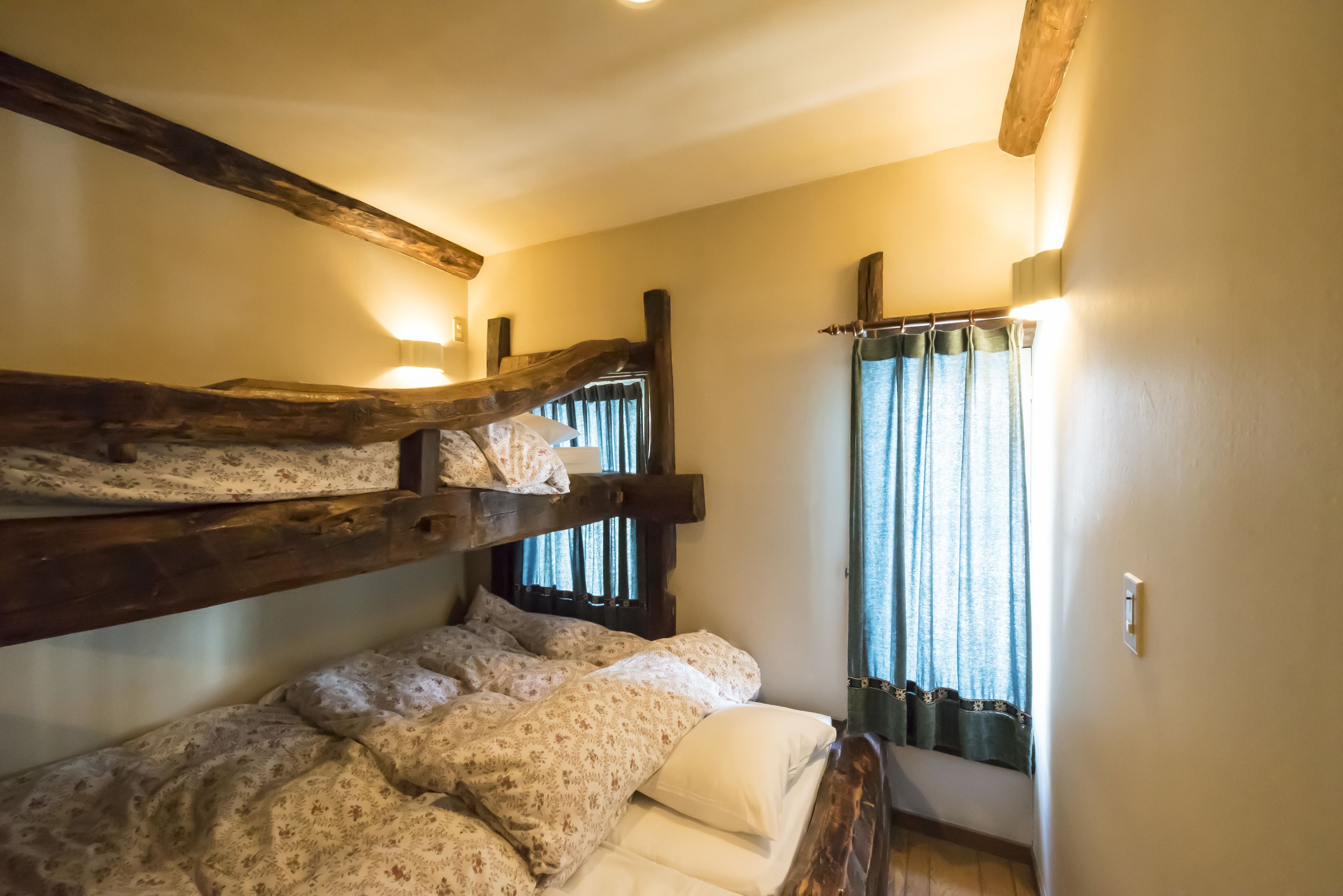 Een slaapkamer met stapelbed, houten balken, een raam met gordijnen en een wandlamp.