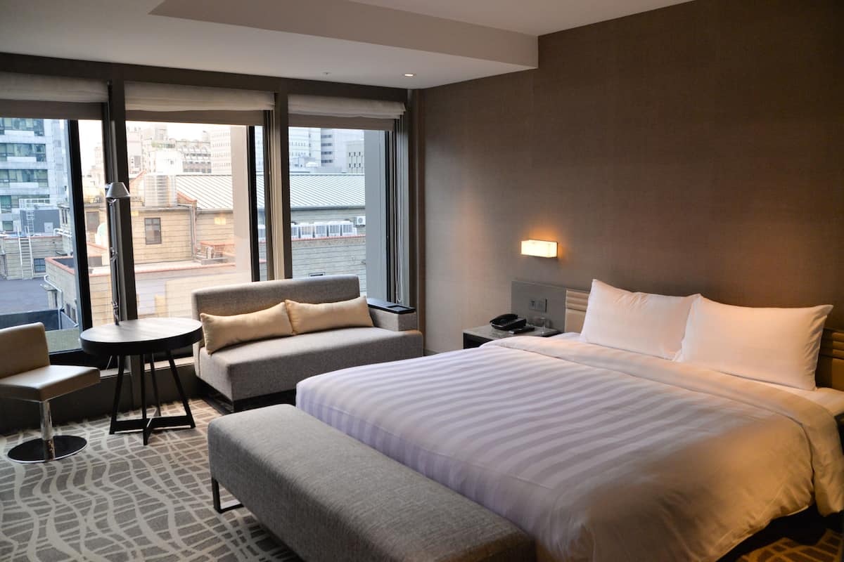 A hotel room with a large bed, a sofa, a small table, and a chair. There is a window with a view of buildings outside.