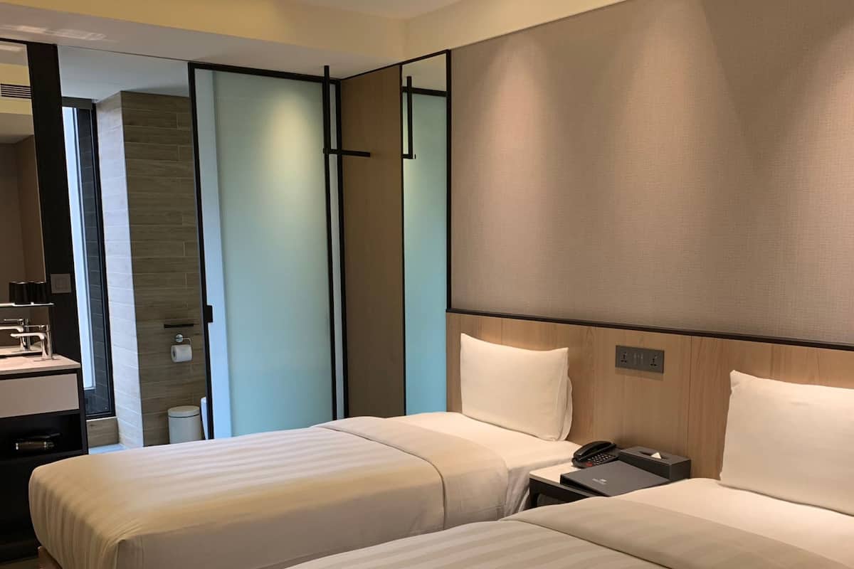 A hotel room with two beds, a bedside table with a phone, and a bathroom visible through a glass door.