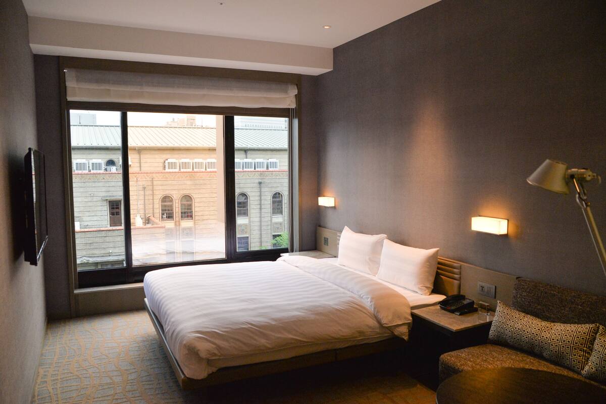 A hotel room with a large bed, bedside tables, a sofa, and a view of buildings through the window.