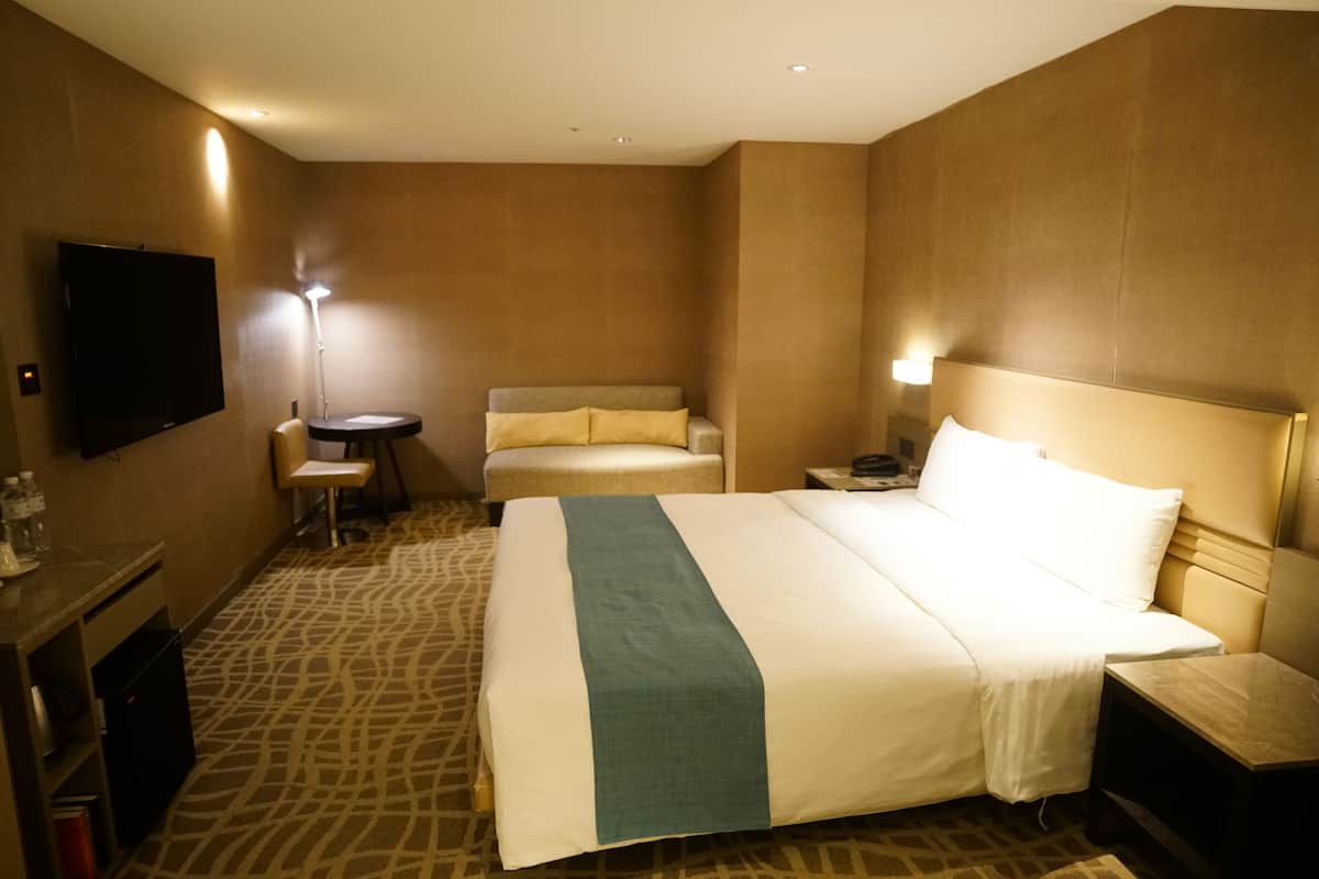A hotel room with a large bed, a bedside table, a chair, a small table, a flat-screen TV, and a wall-mounted TV.