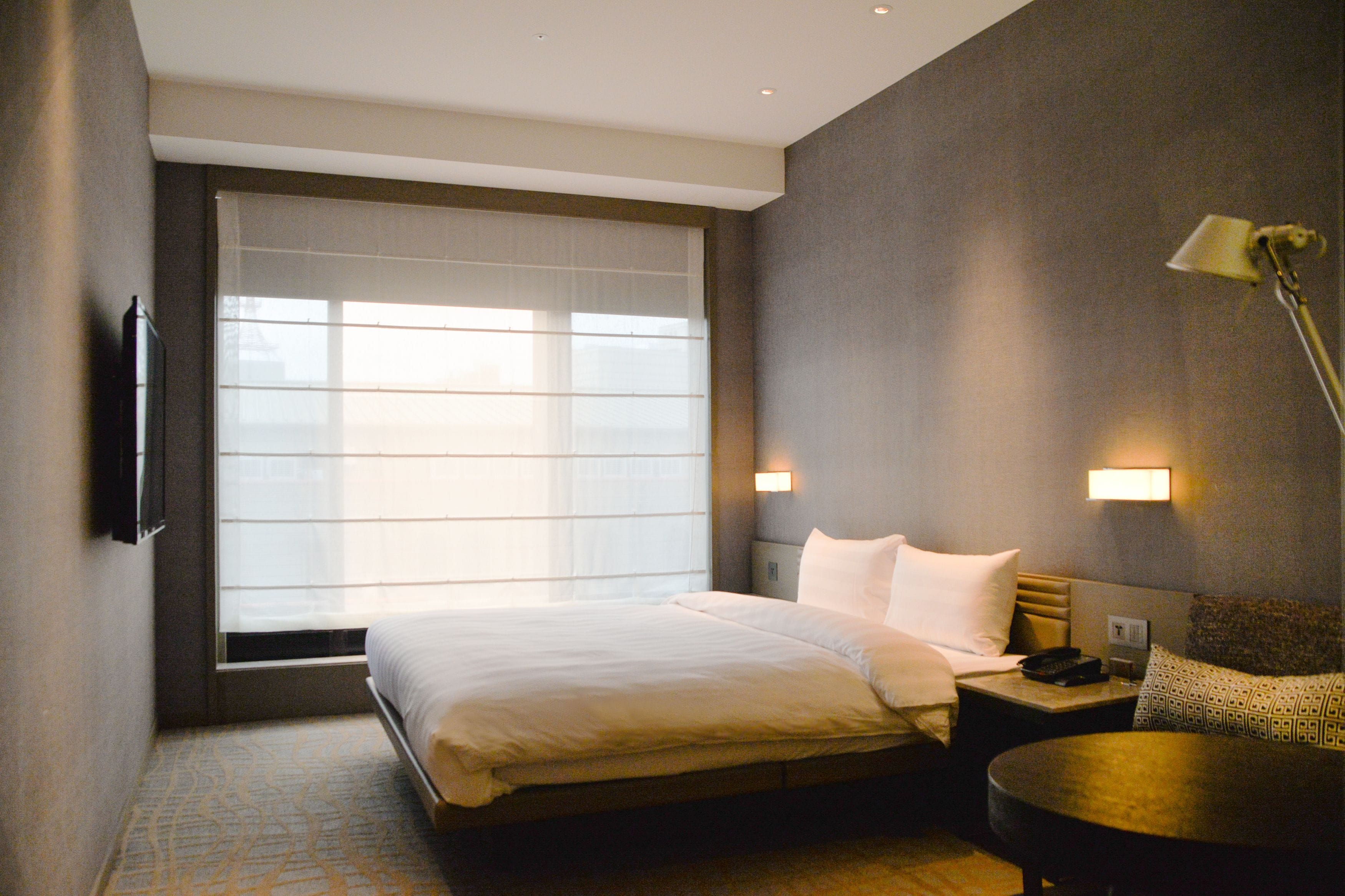A hotel room with a large bed, bedside tables, a desk, and a window with blinds.