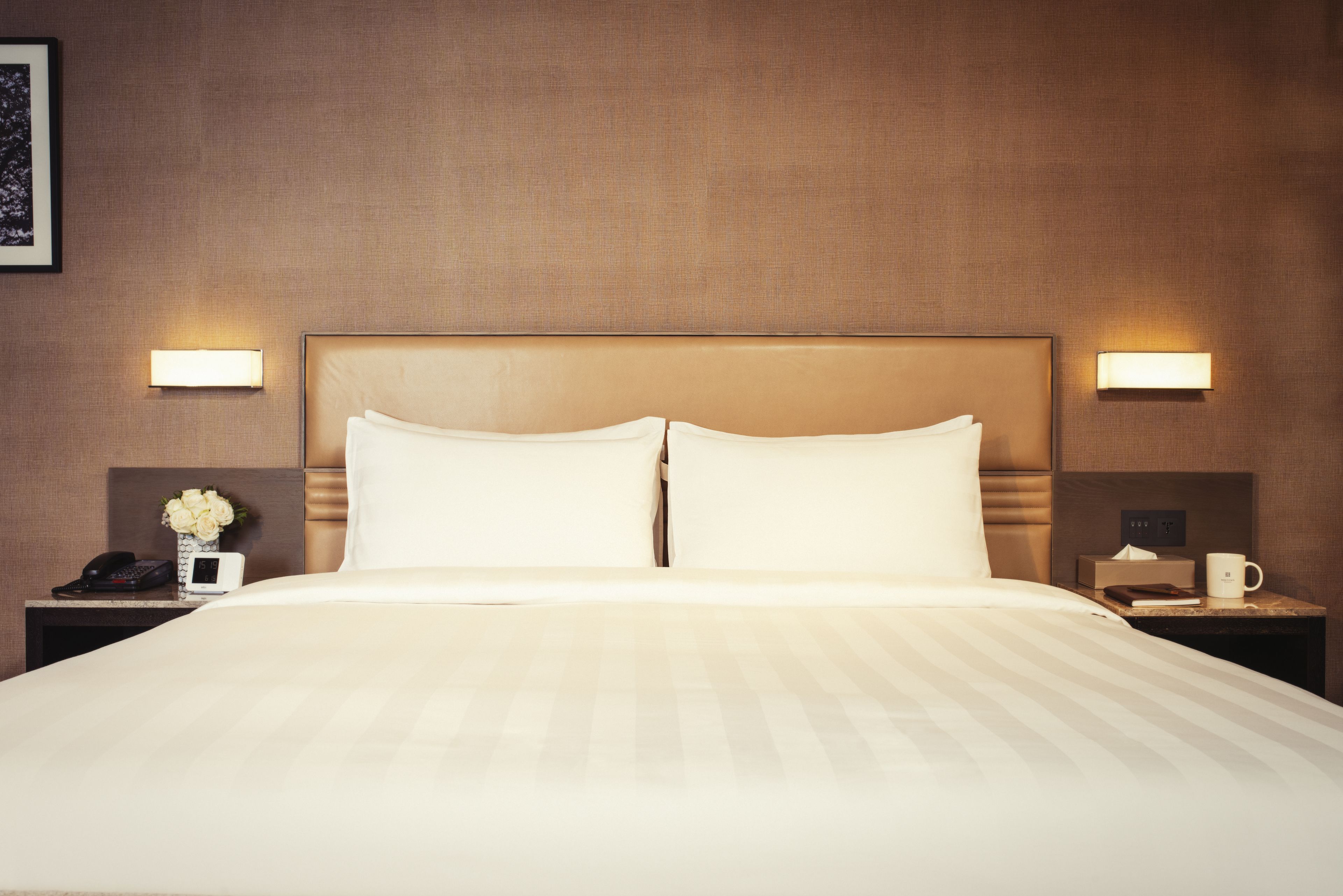 A hotel room with a large bed, two bedside tables, a nightstand, a telephone, a vase with flowers, and two wall-mounted lamps.