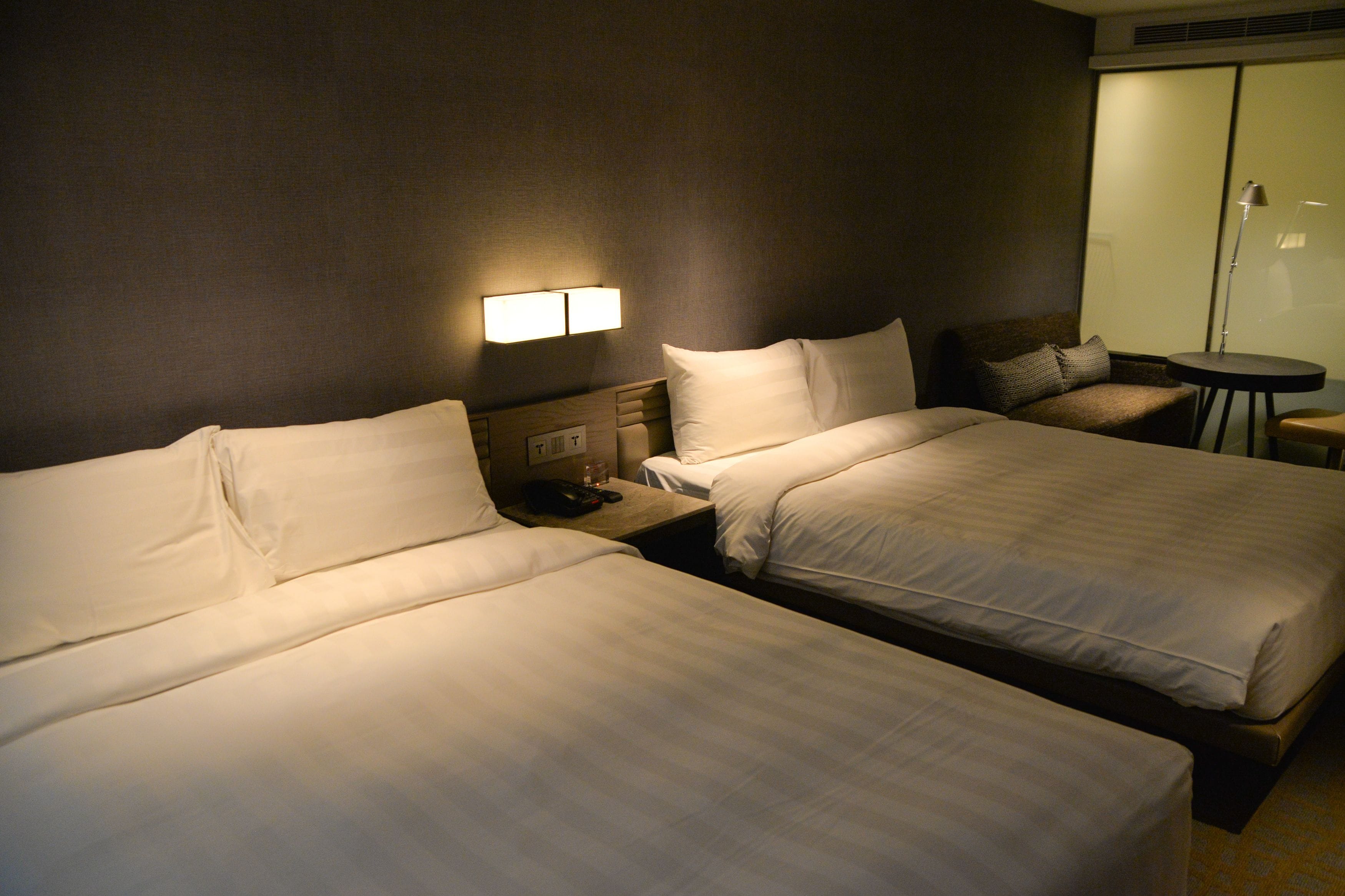 A hotel room with two beds, a bedside table with a lamp, and a small table with a chair.