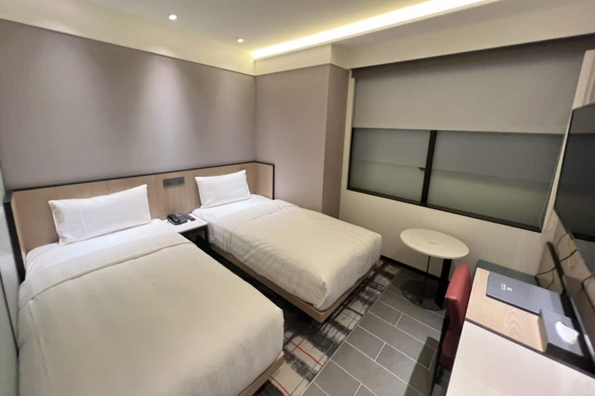 A hotel room with two beds, a small round table, a desk with a computer, and a large window with blinds.