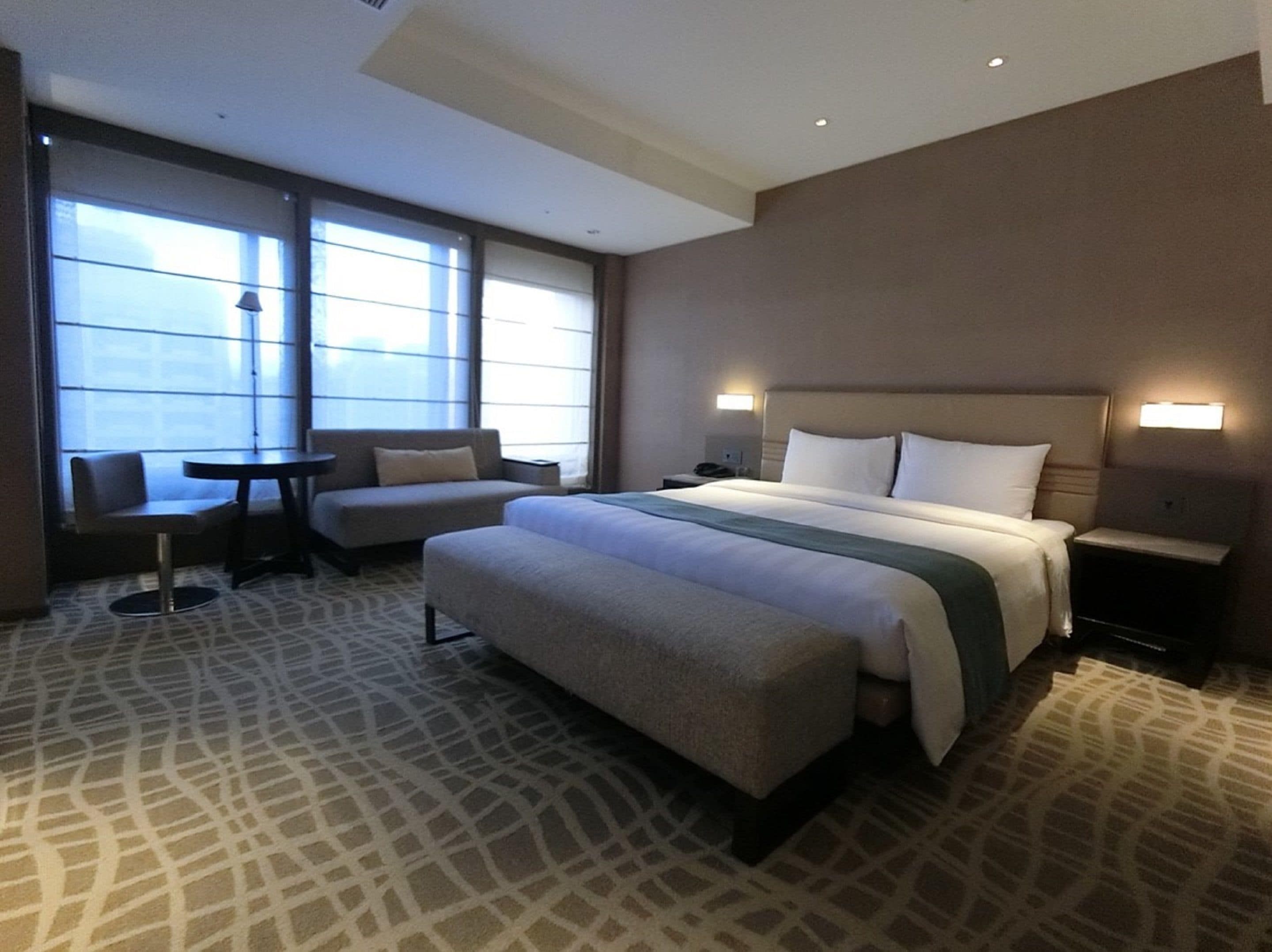 A hotel room with a large bed, a sofa, a small table, and a chair. There are two bedside tables with lamps.