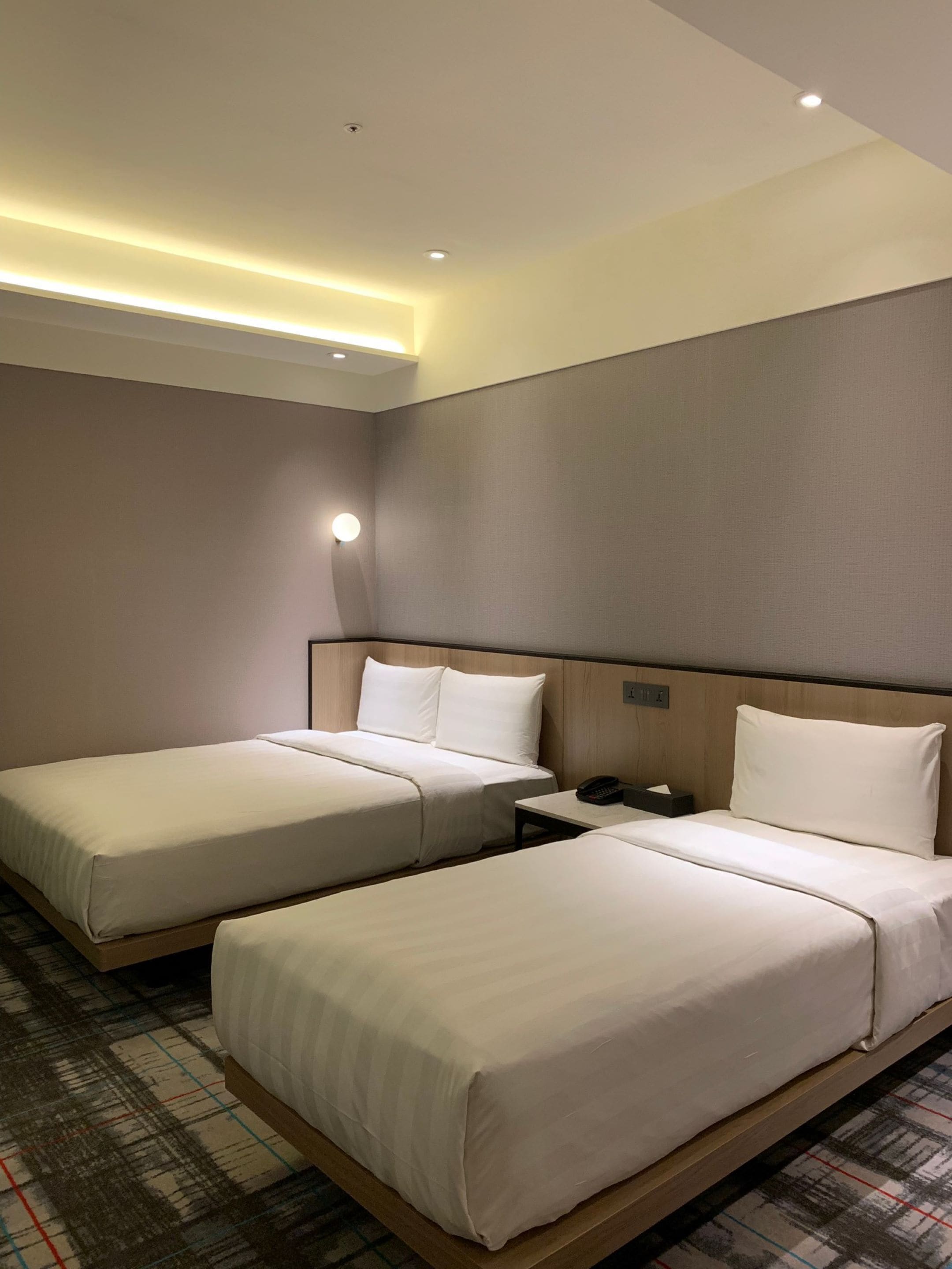 A hotel room with two single beds, a nightstand, a telephone, and a wall-mounted light.