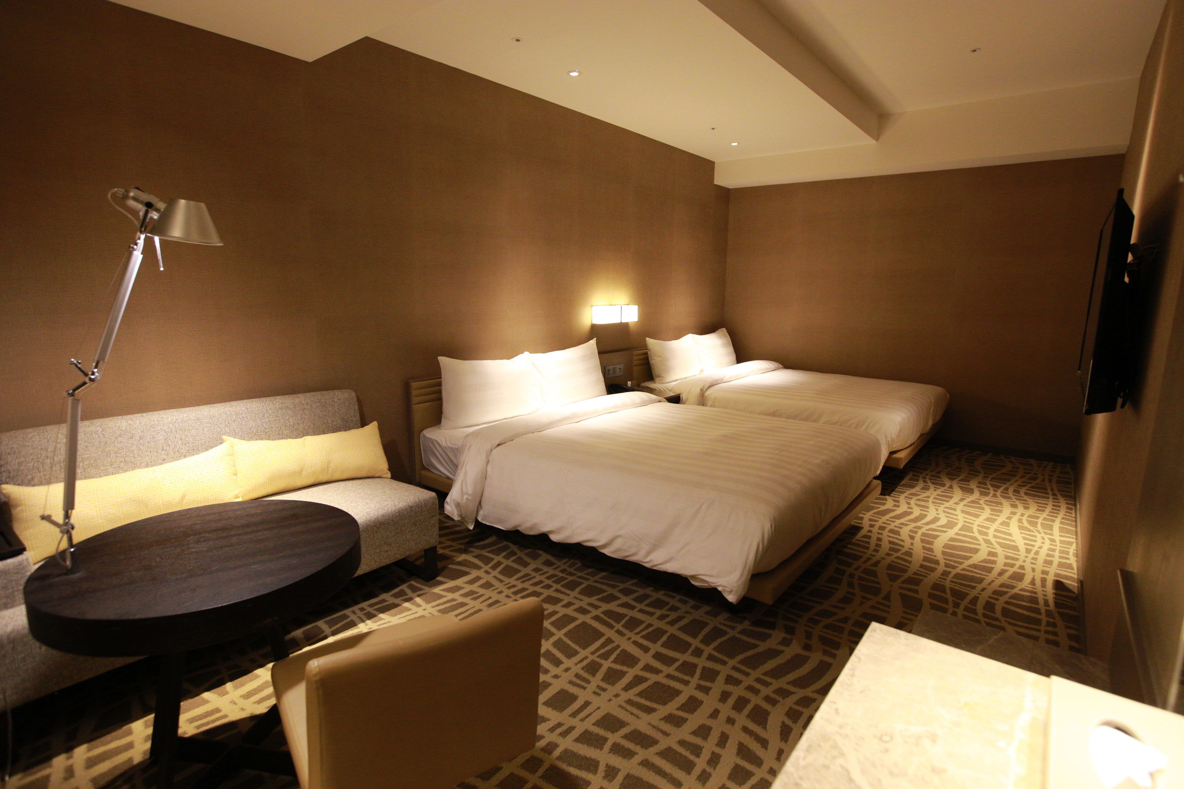 A hotel room with a sofa, two beds, a table, and a lamp.