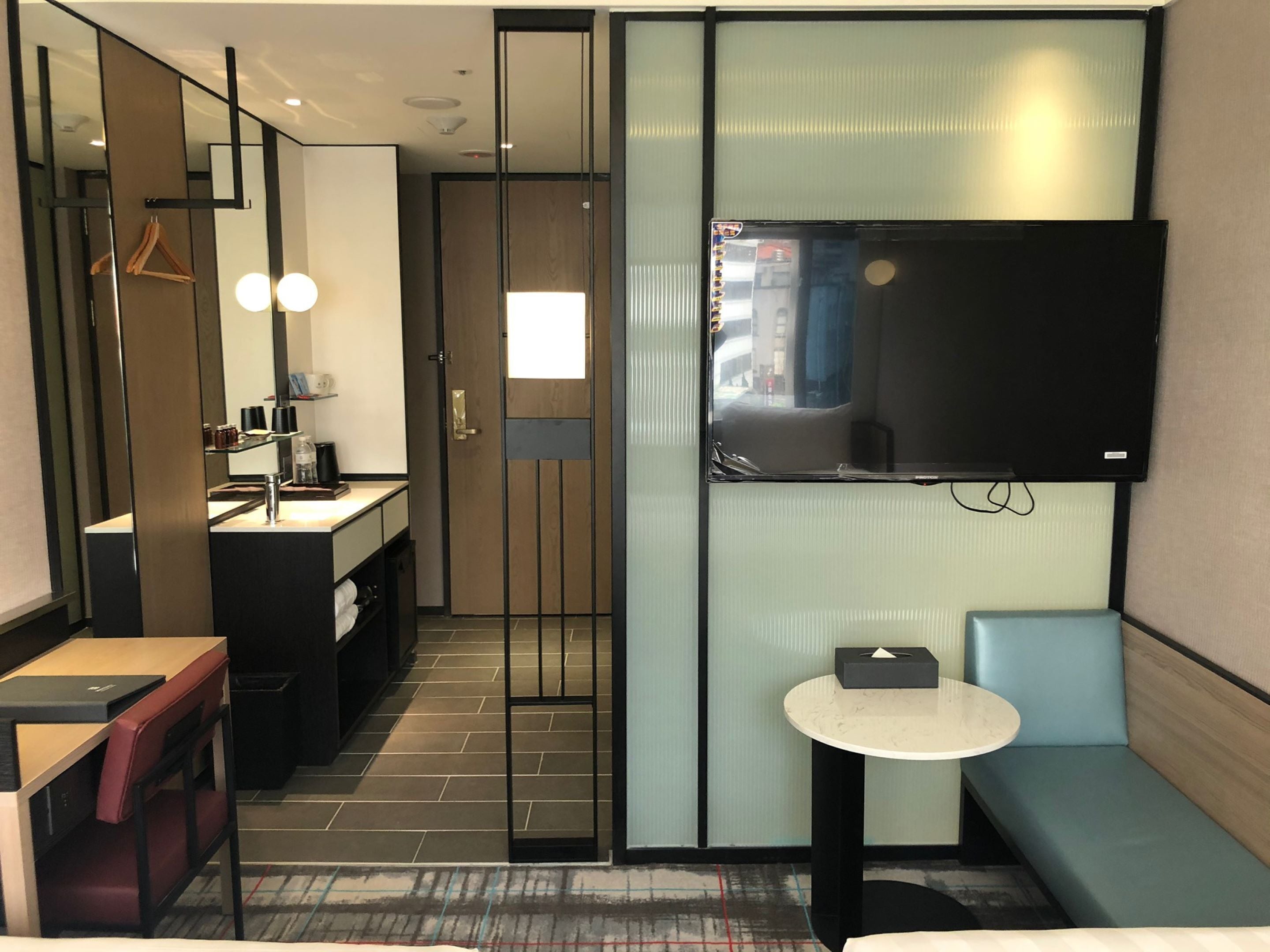 A modern hotel room with a large mirror, a flat-screen TV mounted on the wall, a small round table, a sofa, and a desk with a chair.