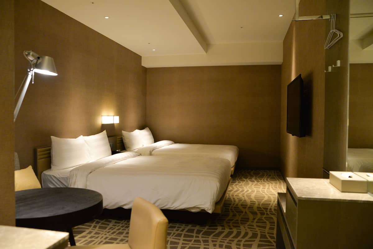 A hotel room with two beds, a desk, a chair, a lamp, a TV, and a patterned floor.
